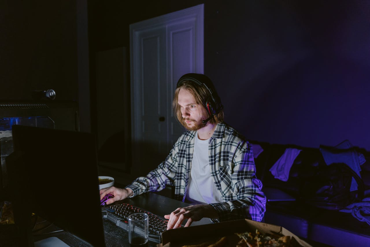 Services-01 A man wearing headphones plays video games on a computer in a dark room, illuminated by screen light.